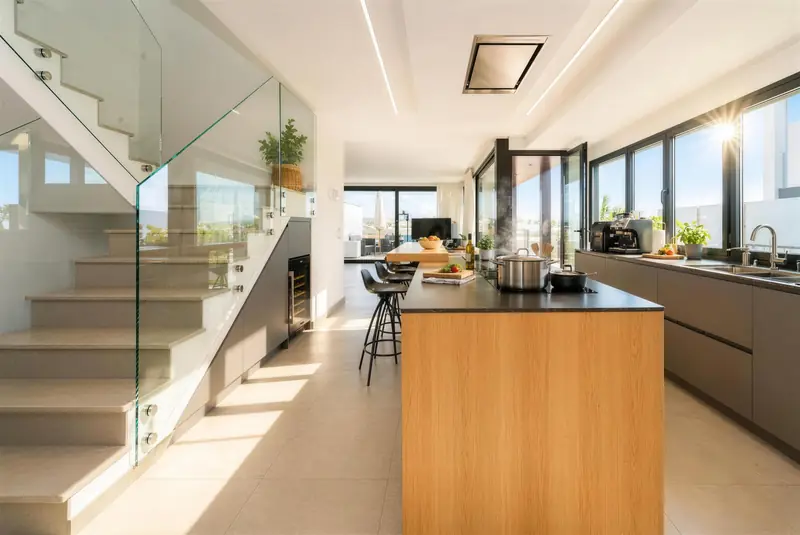Cocina con isla de madera y taburetes negros, escalera de cristal y salón con salida a la terraza